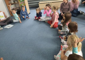 Dzieci siedzą na dywanie i biorą udział w prelekcji