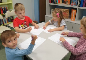 Dzieci stojąc przy stolikach, rysują kredkami na kartonach