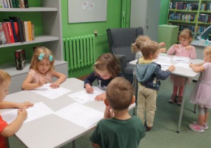 Dzieci stojąc przy stolikach, rysują kredkami na kartonach