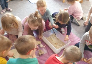 Dzieci przy pomocy pędzelka wyszukują plastikowe fragmenty szkieletów dinozaurów schowane w piasku w kuwetach