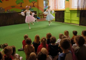 Dzieci siedzą na dywanie i ławczkach oglądając przedstawienie baletowe